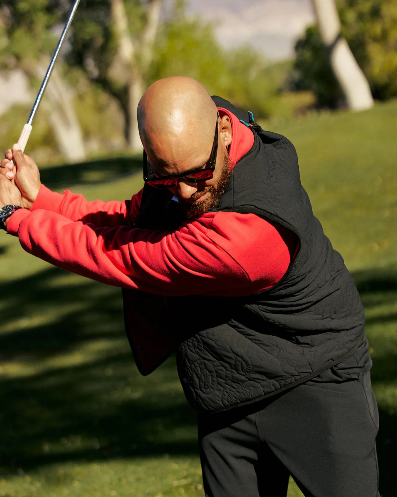 Golfer mid-swing in a black quilted vest over a red long-sleeve top